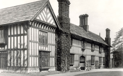 Black and white image of a historic timber-framed house with ivy-covered walls and a large chimney.