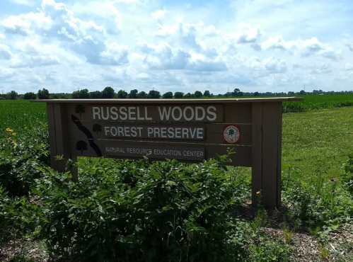 Sign for Russell Woods Forest Preserve, surrounded by green fields and a cloudy sky. Natural resources education center.