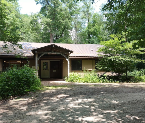 A rustic building surrounded by trees and greenery, with a gravel path leading to the entrance.