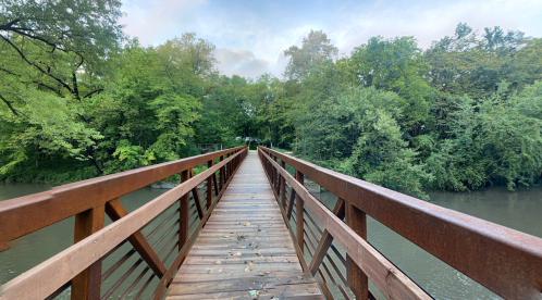 A wooden bridge stretches over a calm river, surrounded by lush green trees and a cloudy sky.