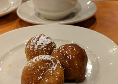 Three golden-brown dough balls dusted with powdered sugar on a white plate, with a cup in the background.