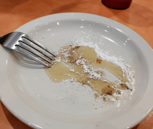 A nearly empty plate with remnants of syrup, powdered sugar, and a fork resting on the edge.