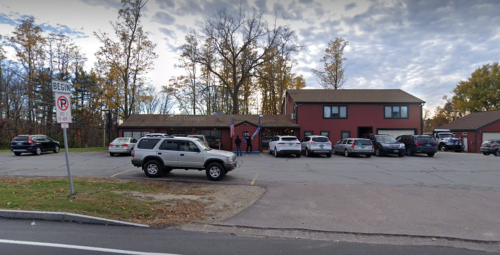 A parking lot with several cars outside a two-story red building surrounded by trees under a cloudy sky.
