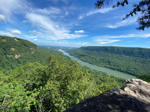 A scenic view of a river winding through lush green hills under a blue sky with scattered clouds.