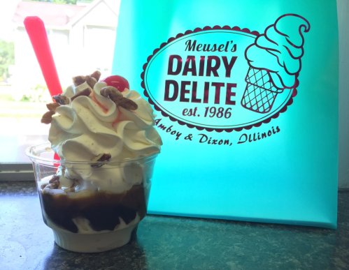 A cup of ice cream topped with nuts and a cherry, next to a teal bag with "Meusel's Dairy Delite" logo.