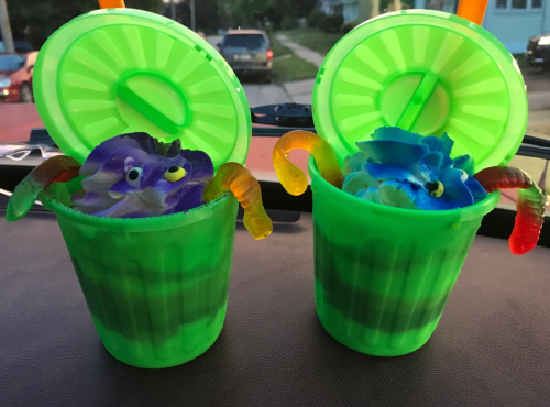 Two green cups filled with colorful whipped cream, gummy worms, and blue decorations, sitting on a car dashboard.