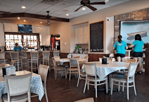 A restaurant interior with tables, a bar area, and staff preparing for service. Light decor and a casual atmosphere.