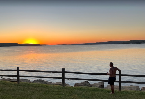 A person stands by a wooden fence, watching a vibrant sunset over calm water.