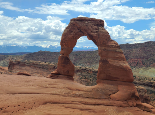 A natural stone arch stands against a backdrop of mountains and cloudy skies in a desert landscape.