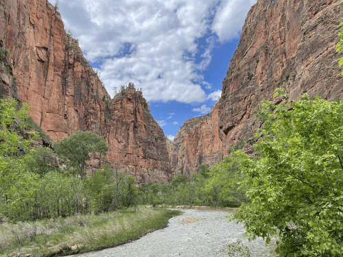A scenic view of a canyon with towering red rock cliffs, a river, and lush green trees under a partly cloudy sky.