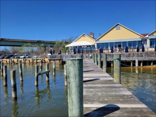 A wooden pier extends over calm water, leading to a yellow building with outdoor seating under blue skies.