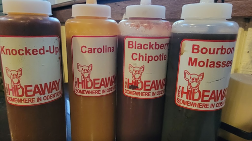 Four condiment bottles labeled "Knocked-Up," "Carolina," "Blackberry Chipotle," and "Bourbon Molasses" on a table.