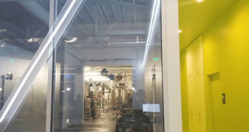 A modern brewery interior with stainless steel tanks visible through large glass windows and a bright yellow wall.