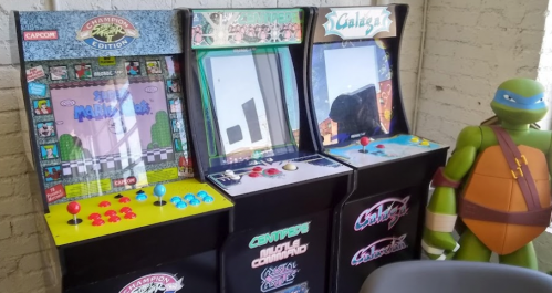 Three vintage arcade machines with colorful controls, alongside a green turtle character figure.