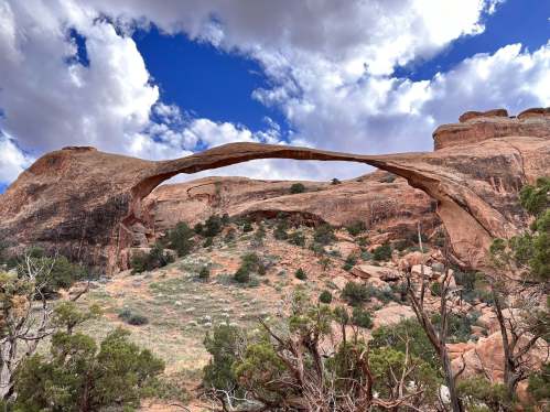Natural stone arch rises above a rugged landscape, framed by blue skies and scattered clouds. Green shrubs in the foreground.