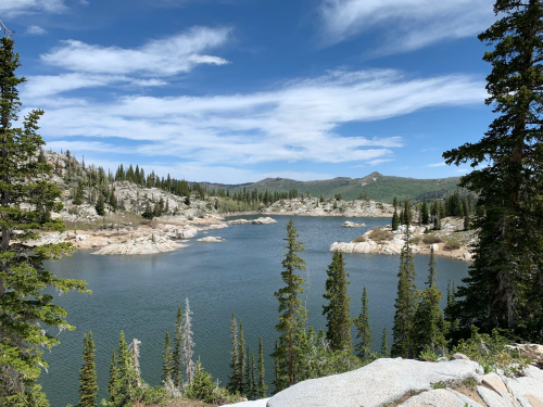 A serene lake surrounded by rocky shores and tall pine trees under a blue sky with scattered clouds.