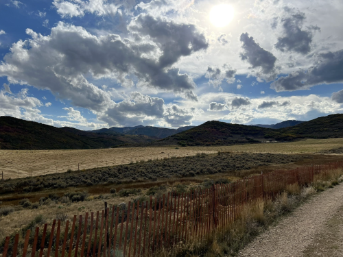 A scenic landscape featuring rolling hills, a cloudy sky, and a sunlit field, with a dirt path and wooden fence in the foreground.