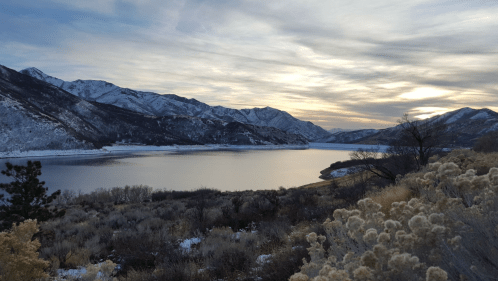 A serene lake surrounded by snow-covered mountains under a cloudy sky at sunset.