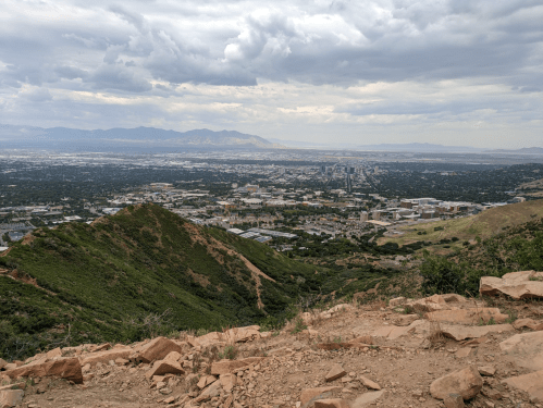 A panoramic view of a city nestled in a valley, surrounded by mountains under a cloudy sky.