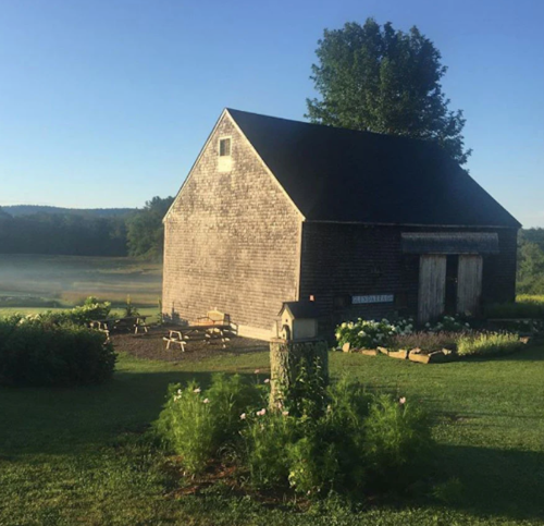 A rustic barn surrounded by greenery and flowers, with a clear blue sky and morning mist in the background.