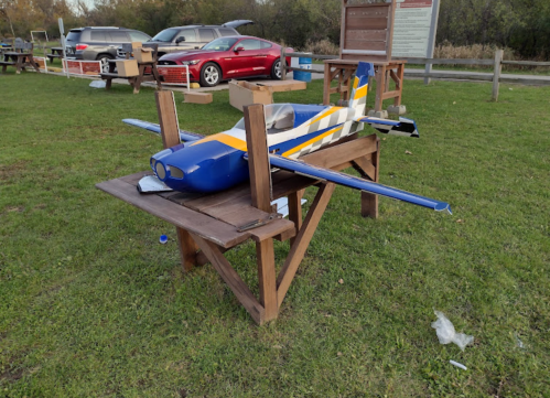 A model airplane with blue and yellow stripes sits on a wooden table in a grassy area, surrounded by parked cars.