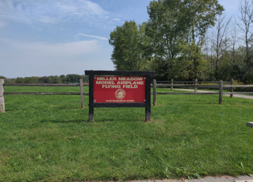 Sign for Miller Meadow Model Airplane Flying Field, surrounded by grass and trees under a clear sky.