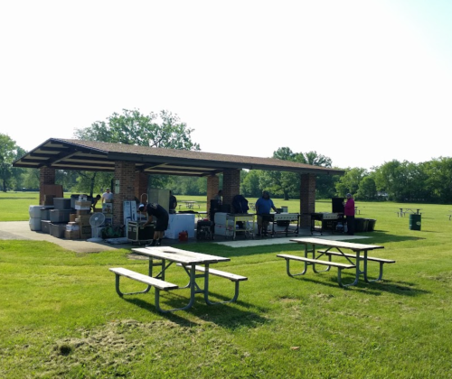 A park pavilion with people setting up tables and grills, surrounded by green grass and trees on a sunny day.
