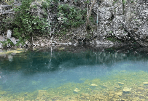A serene, clear blue-green pool surrounded by rocky terrain and lush greenery. Reflections shimmer on the water's surface.