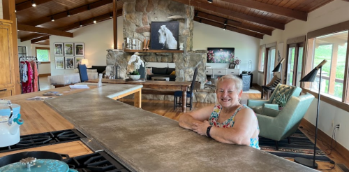 A smiling woman sits at a kitchen island in a spacious, modern home with a stone fireplace and large windows.