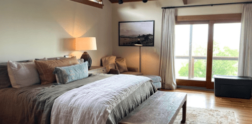 Cozy bedroom with a large bed, wooden furniture, and a view of greenery through a window. Soft lighting creates a warm atmosphere.