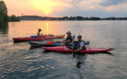 Three people in kayaks fishing at sunset on a calm lake, with trees and a distant shoreline in the background.