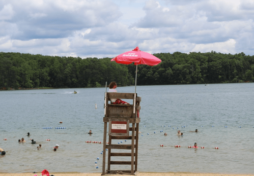 A lifeguard sits on a tall chair by a lake, watching swimmers under a red umbrella, with trees in the background.