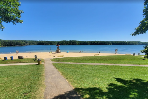 A sunny beach scene with a sandy shore, lifeguard stands, and calm water surrounded by green trees.