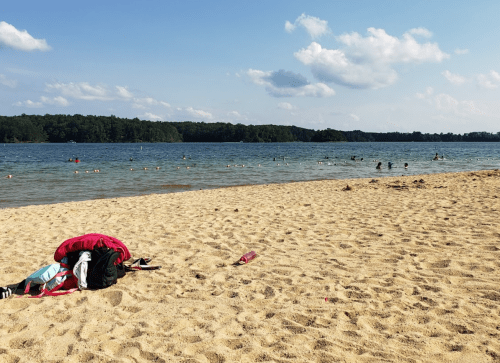 A sandy beach with a calm lake, people swimming in the distance, and a pile of clothes on the shore.