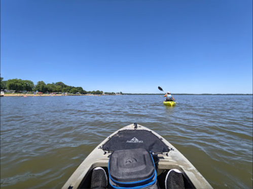 A view from a kayak on a calm lake, with another kayaker in the distance and a clear blue sky above.