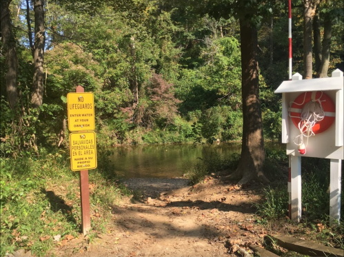 A path leads to a calm water area, marked with signs warning of no lifeguards and safety instructions.