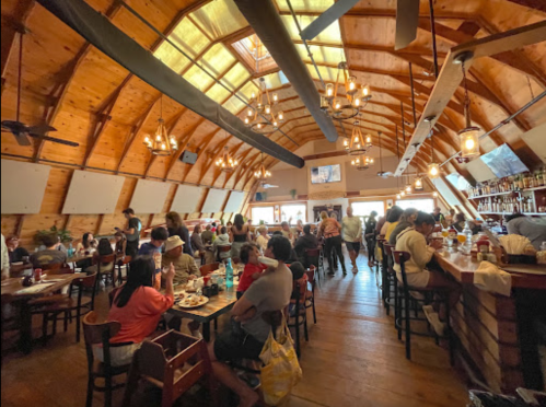 A bustling restaurant interior with wooden beams, patrons dining, and a bar area filled with drinks and food.