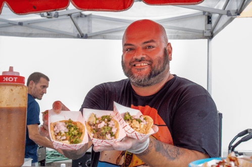 A smiling vendor holds three tacos in paper trays, with a food stand in the background.