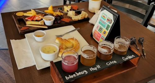 A wooden table with two platters: one with cheese, fruits, and snacks, and another with beer samples and a pretzel.