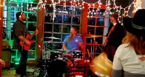 A lively band performs in a cozy venue decorated with string lights, with a drummer and guitarists engaging the audience.