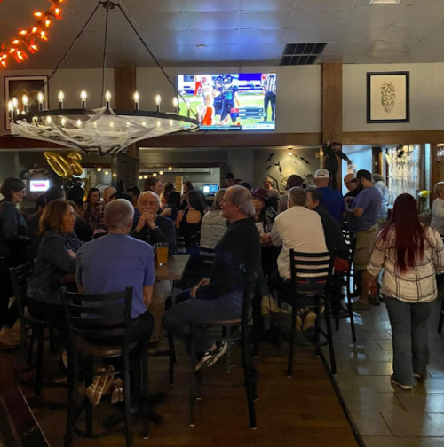 A lively bar scene with patrons gathered around tables, watching a game on a TV, and enjoying drinks.