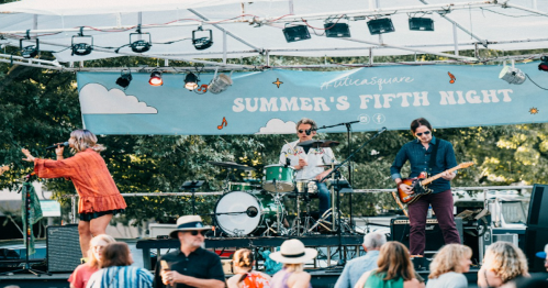 A band performs on stage at a summer event, with a crowd enjoying the music under a clear sky.