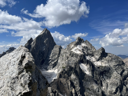 A panoramic view of rugged mountain peaks under a blue sky with scattered clouds. Snow patches visible on the slopes.