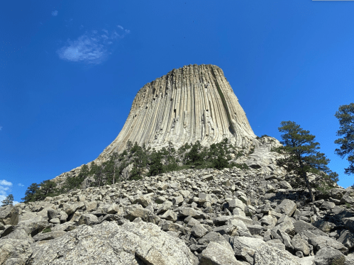 A towering rock formation rises against a blue sky, surrounded by trees and rocky terrain.