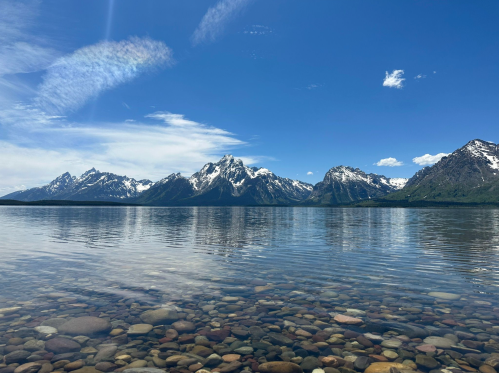 A serene lake reflects snow-capped mountains under a clear blue sky, with colorful pebbles visible beneath the water.