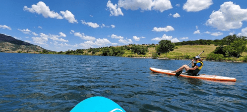 A person paddles on a stand-up paddleboard in a lake surrounded by green hills and a blue sky with fluffy clouds.