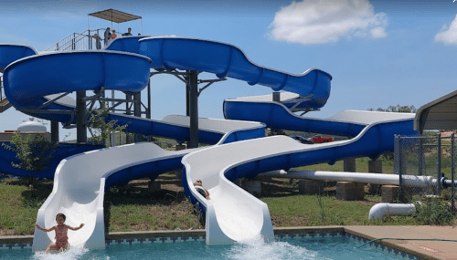 A water park with blue and white slides, a child splashing into the pool, and people on the upper platform.