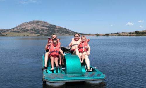 Four people, including two children, enjoy a pedal boat ride on a lake with a mountain in the background.