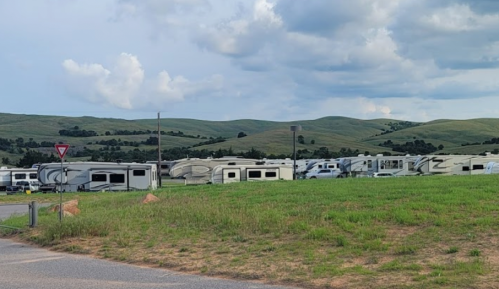 A row of RVs parked in a grassy area with rolling hills and cloudy skies in the background.
