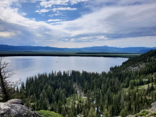 A serene landscape featuring a calm lake surrounded by lush forests and distant mountains under a cloudy sky.